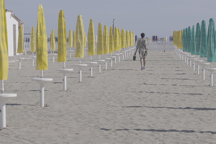 scena tratta da un documentario: una persona che cammina sulla spiaggia tra gli ombrelloni chiusi